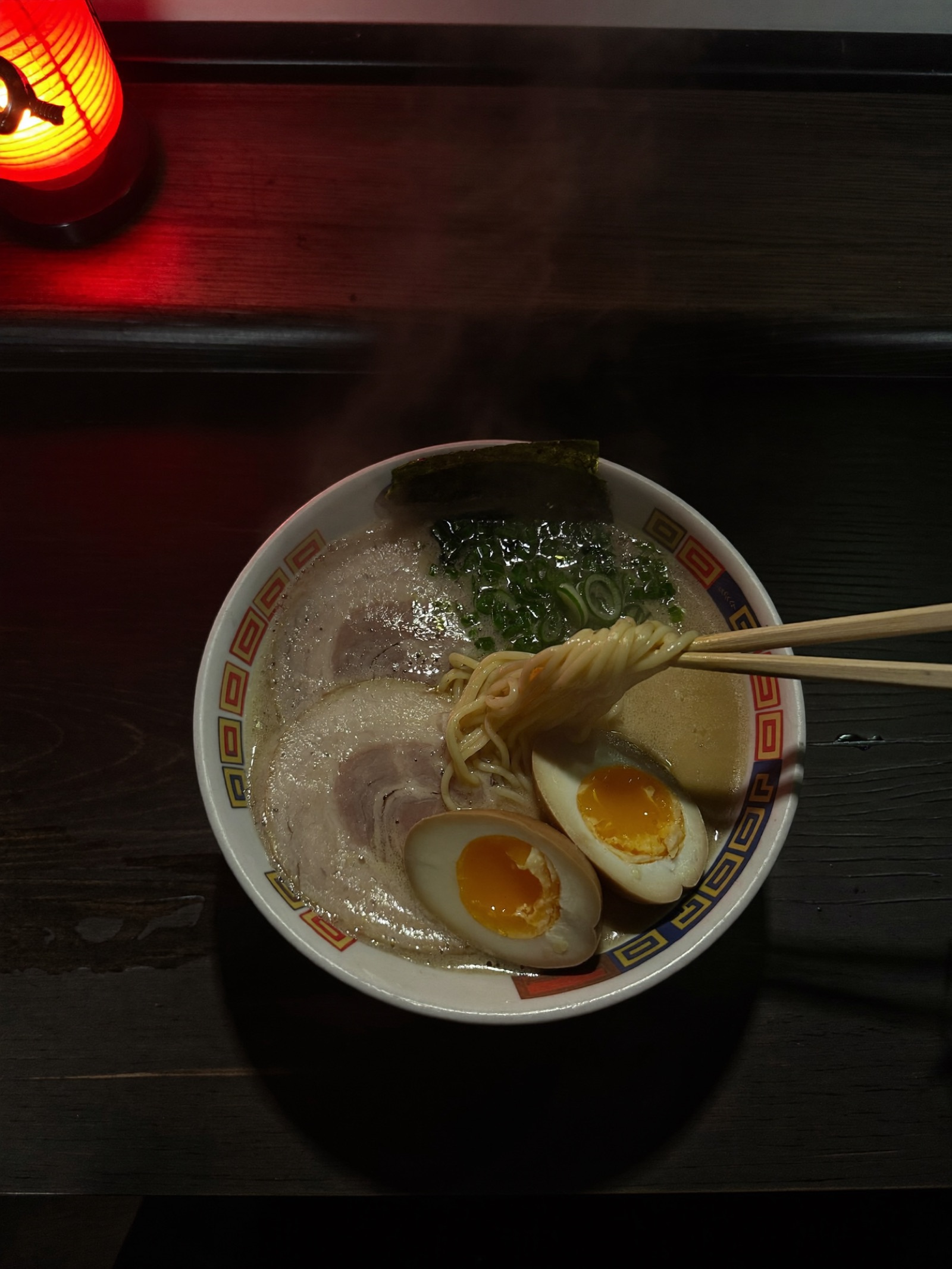 A bowl of tonkotsu ramen at the Menko counter, cloud-white broth and chashu lit by the red glow of a paper lantern, steam rising into the dark